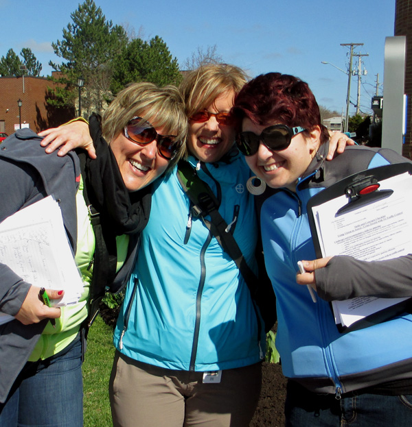 Photo shows Josee, Dominique and Denise with arms around each other, laughing and smiling at the camera. Two of them are holding clipboards.